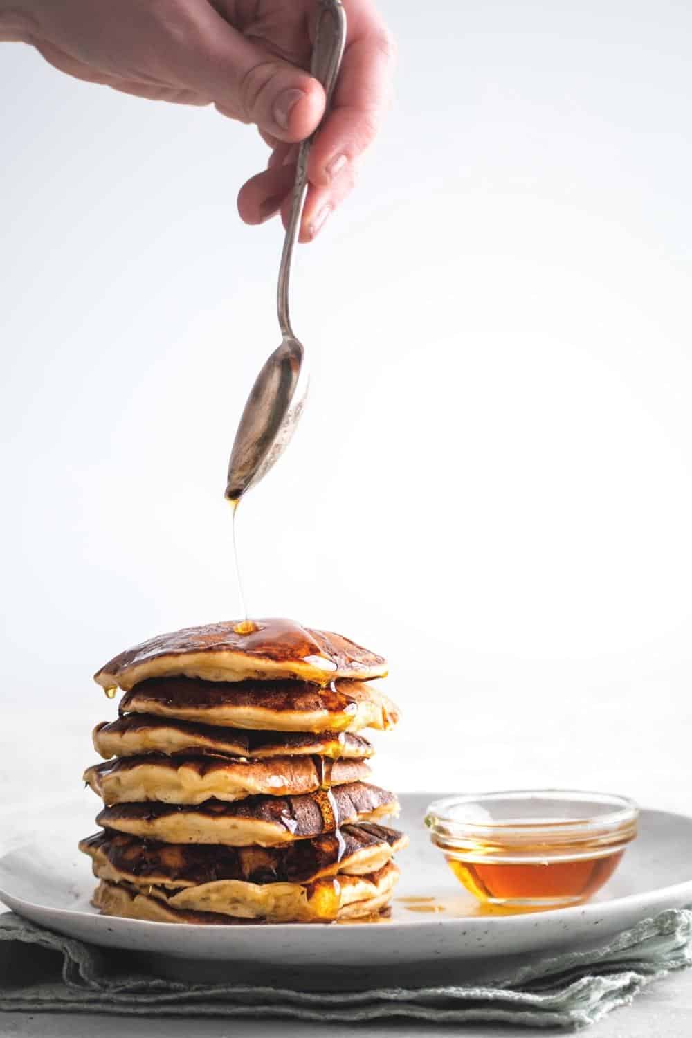A hand holding a spoon drizzling syrup on top of a stack of seven pancakes on a white plate.