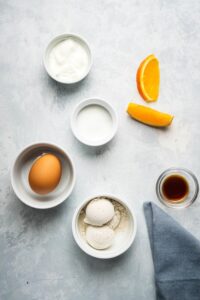 A bowl of oat flour, a bowl with an egg in it, a small bowl with vanilla extract in it, small bowl with erythritol in it, and a small bowl Greek yogurt all on a gray counter.