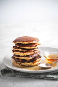 A stack of seven pancakes on a white play with a spoon and part of a bowl of honey on it.