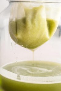 Celery juice in a cheese cloth that is inside a glass jar that has celery juice in it.