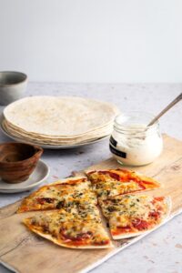 A tortilla pizza on top of a wooden cutting board. There is a glass bowl of ranch next to it and behind it is a bunch of flour tortillas on a plate.