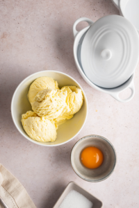 A few scoops of ice cream in a bowl, and egg yolk in a bowl, and sugar and part of a ball on a white counter.