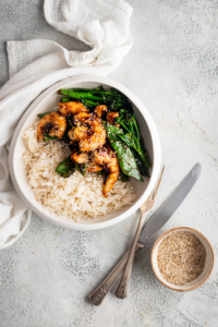 Rice, shrimp, snow peas, and broccoli in a white bowl on a gray counter. Next to it is a fork and knife in a bowl sesame seeds.