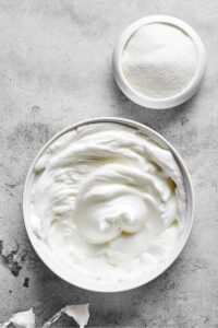 Whipped egg whites in a bowl with sugar and a bowl behind it both on a gray counter.