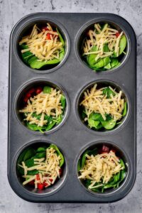 Cheddar cheese on top of spinach on top of diced red pepper in six slots of a muffin tin.