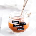 A glass jar filled with sugar-free caramel syrup. There's a spoon in the jar and the jar is on a serving square on the counter.