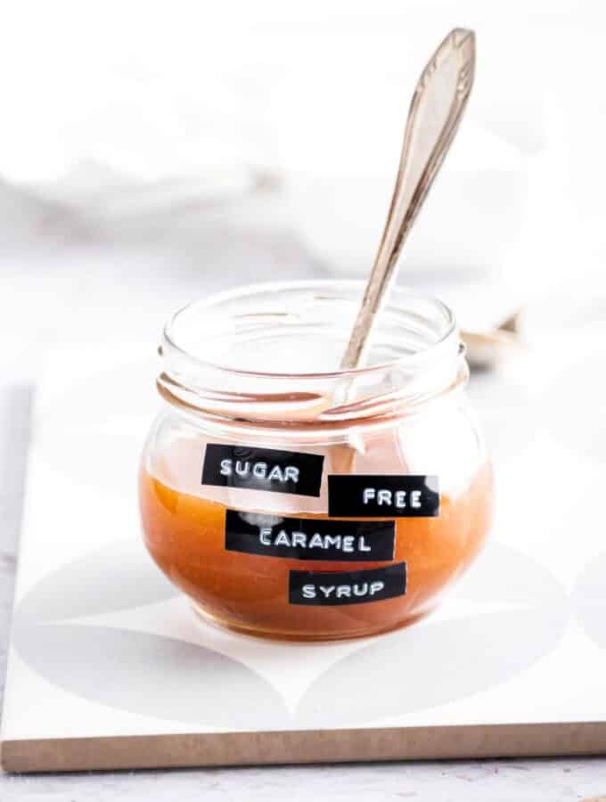 A glass jar filled with sugar-free caramel syrup. There's a spoon in the jar and the jar is on a serving square on the counter.