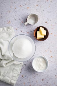 A bowl of granulated sweetener, a bowl of heavy cream, a bowl of cubed butter, in a bowl of salt on a gray counter.