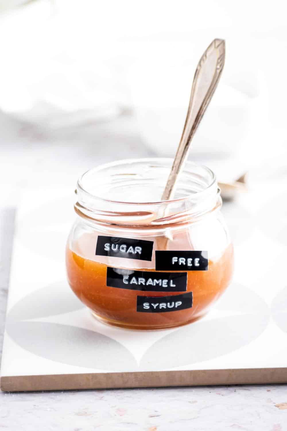 A glass jar filled with sugar-free caramel syrup. There's a spoon in the jar and the jar is on a serving square on the counter.