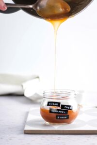 A spoon pouring caramel sauce from apart into a glass jar that is halfway filled with the caramel sauce. Glass jar has sugar free caramel syrup labeled on it.