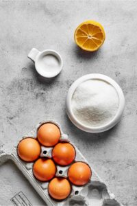 Six eggs in an egg carton, sugar in a bowl, salt in a bowl, and a lemon and a gray counter.