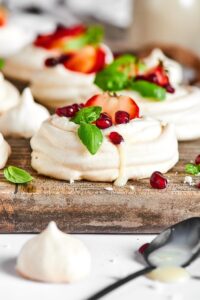 Meringue cookie that has a say strawberry in a few dried pomegranates on top of it on a wooden cutting board.