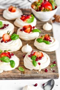 Seven meringue cookies with strawberries and dried pomegranates on top of them on a wooden cutting board.