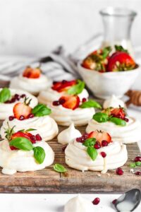 A bunch of meringue cookies on a wooden cutting board. They have strawberries on top and there is a bowl of strawberries behind the cutting board.