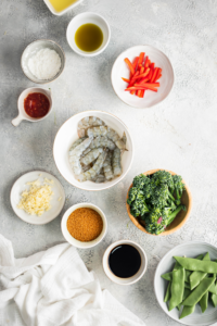 Football snow peas, bowl soy sauce, bowl brown sugar, boil broccoli, bowl shrimp, a bowl of garlic, a bowl of red bell pepper, and a bowl of soy sauce all on a grey counter.