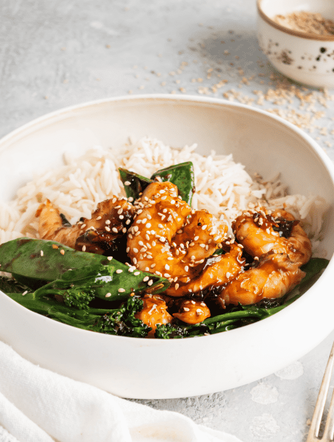 Sesame seeds on top of shrimp, snow peas, broccoli, and white rice in a white bowl.