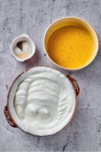 A bowl of whipped egg whites, a bowl of whipped egg yolk, and a bowl of salt pepper garlic and onion powder all on a grey counter.