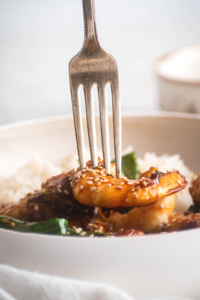 A fork stabbing a piece of shrimp that is on top of some more shrimp and part of a white bowl.