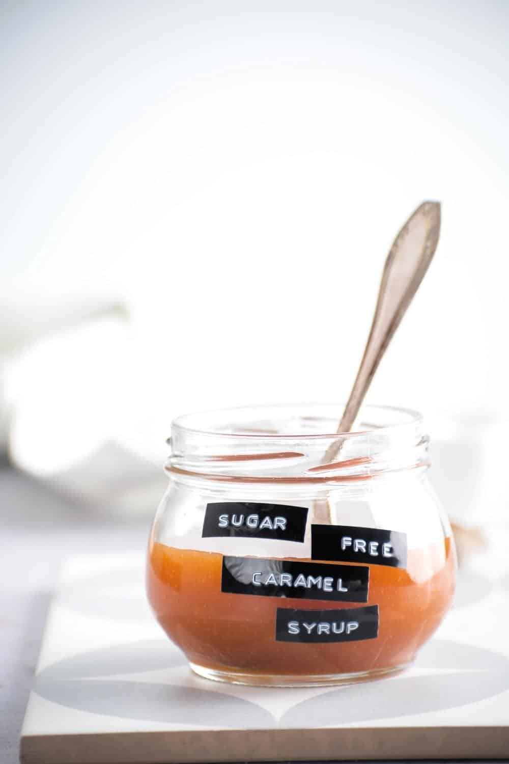 A glass jar halfway filled with caramel sauce. Glass jar has black labels on it that says sugar-free caramel syrup.