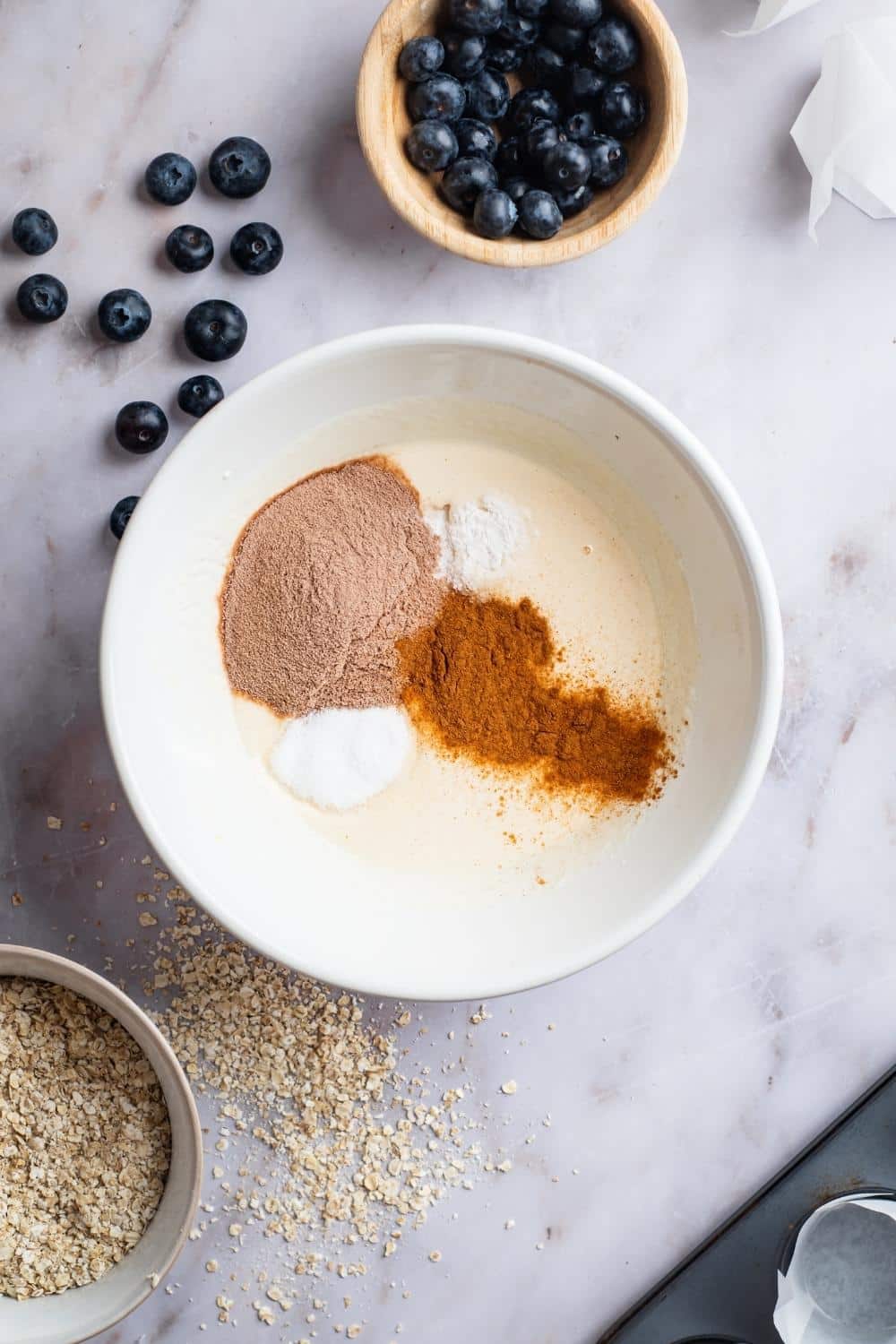 Protein powder, cinnamon, and baking powder in a bowl. There is a cup of blueberries behind it.