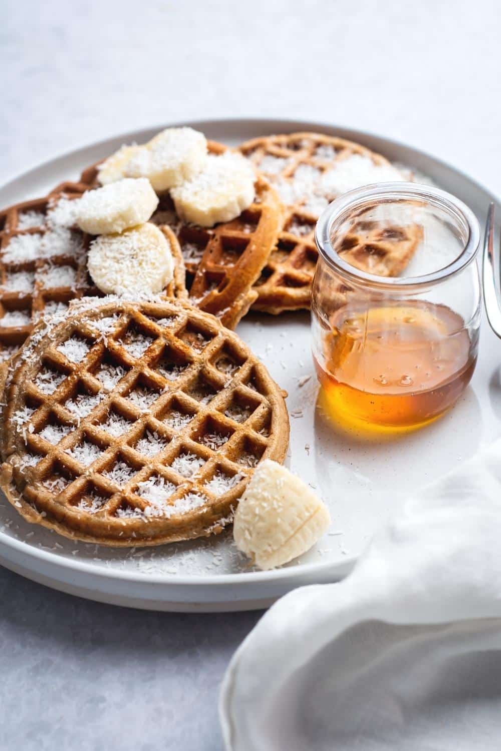 A white plate that has four protein powder waffles on it. There's shredded coconut and sliced bananas on two of the waffles.
