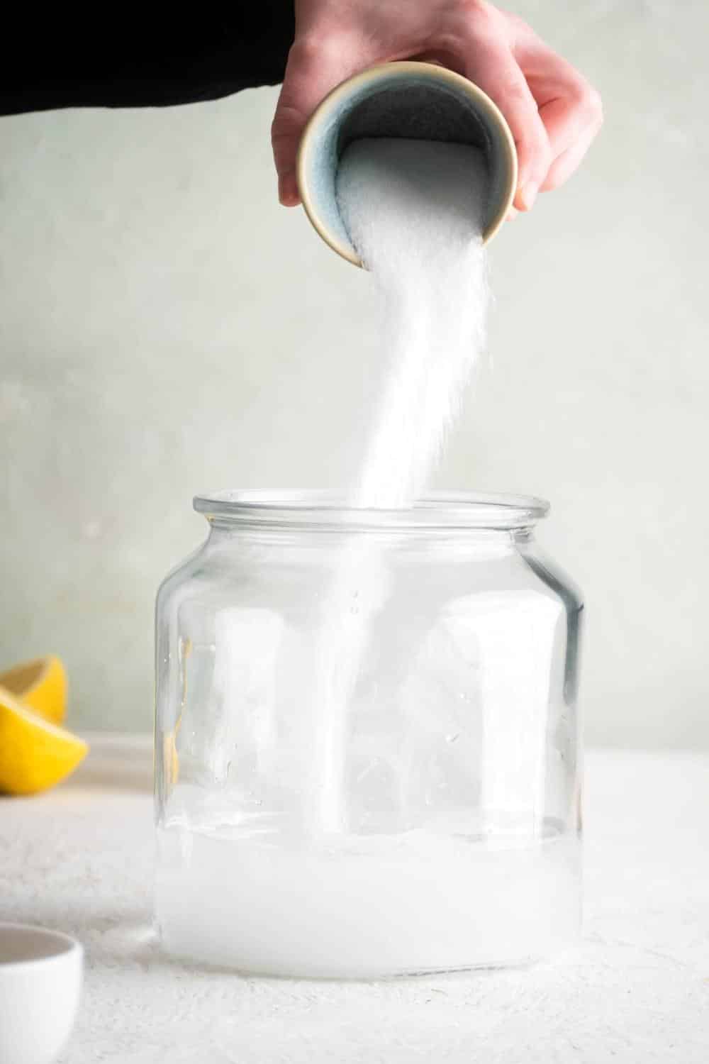 A hand pouring granulated sweetener into a large glass bowl.