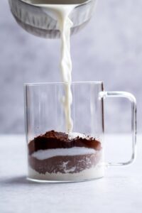 Milk being poured into a glass mug that is filled with cocoa powder, chocolate protein powder, and oat flour.