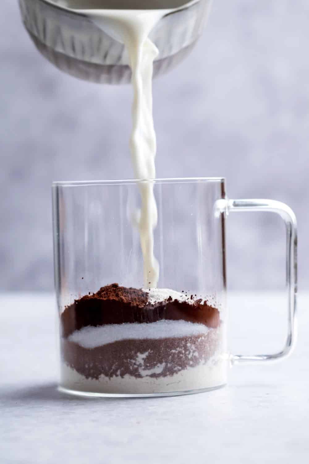 Milk being poured into a glass mug that is filled with cocoa powder, chocolate protein powder, and oat flour.