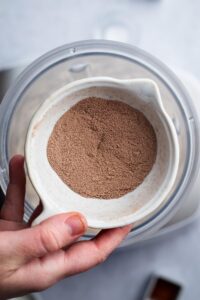 Hand holding a white bowl of chocolate protein powder over a blender.