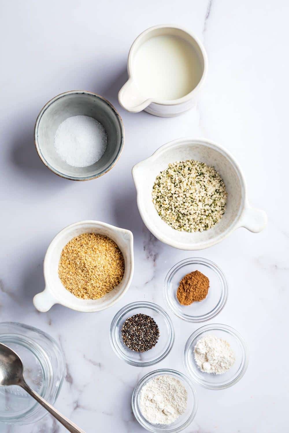 A picture of milk, a bowl of confectioners erythritol, a bowl of ham parts, a bowl of golden flaxseed, a bowl of Chia seeds, bowl cinnamon, a bowl of almond flour, animal of coconut flour all on a white counter.