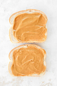 Two slices of bread with peanut butter on them. The slices are on a white table.