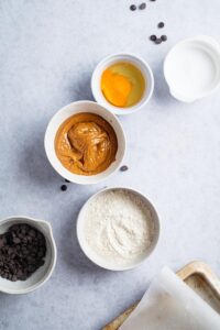 A bowl of peanut butter, a bowl of protein powder, a bowl of egg, and a bowl of chocolate chips all on a white counter.