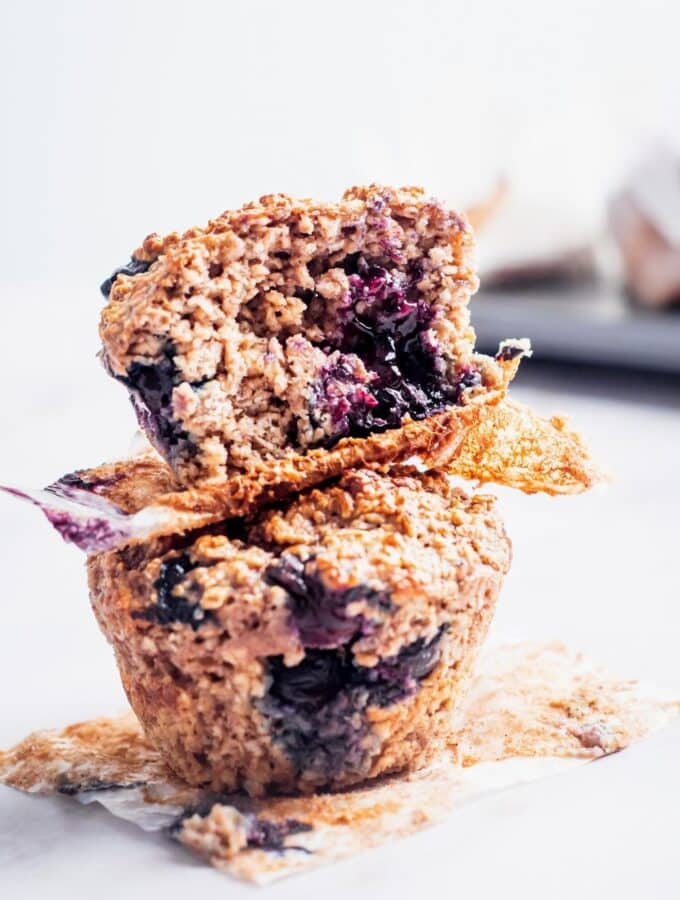 Two blueberry protein powder muffins on top of one another. the top muffin has a bite out of the front.