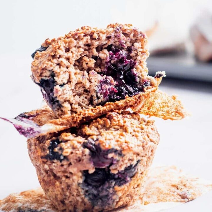 Two blueberry protein powder muffins on top of one another. the top muffin has a bite out of the front.