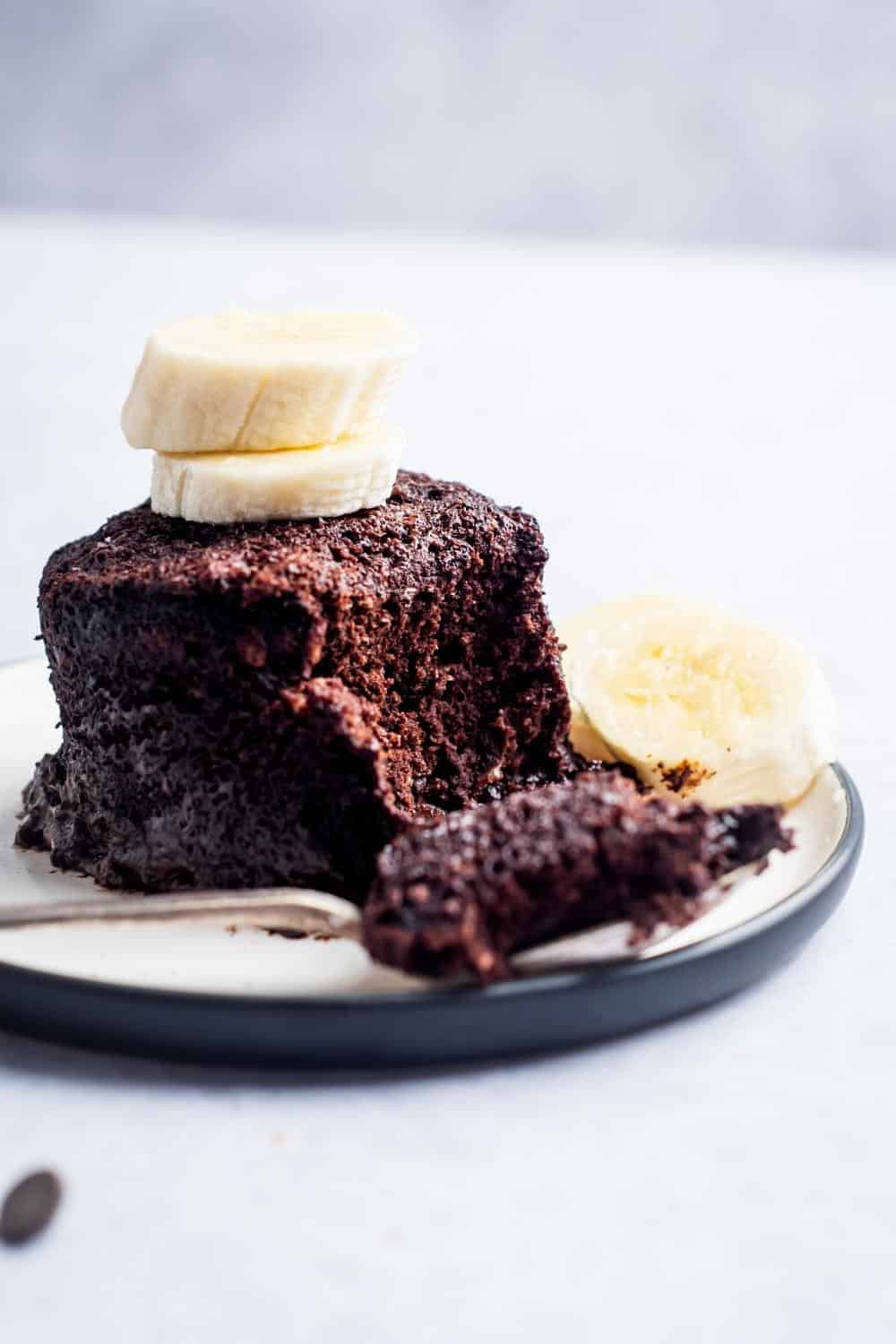 A chocolate protein mug cake and a white plate with a piece of the mug cake taken out of the front of it.