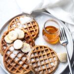For protein waffles with some shredded coconut and sliced bananas on top on a white plate with a fork in a jar maple syrup.