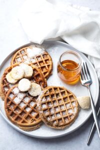 For protein waffles with some shredded coconut and sliced bananas on top on a white plate with a fork in a jar maple syrup.