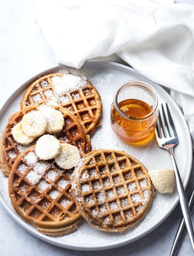 For protein waffles with some shredded coconut and sliced bananas on top on a white plate with a fork in a jar maple syrup.