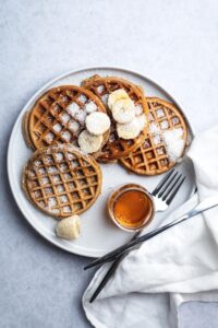 Four protein waffles overlap in one another with some shredded coconut and some sliced bananas on top. The waffles are on a plate with a fork and a glass jar of syrup.