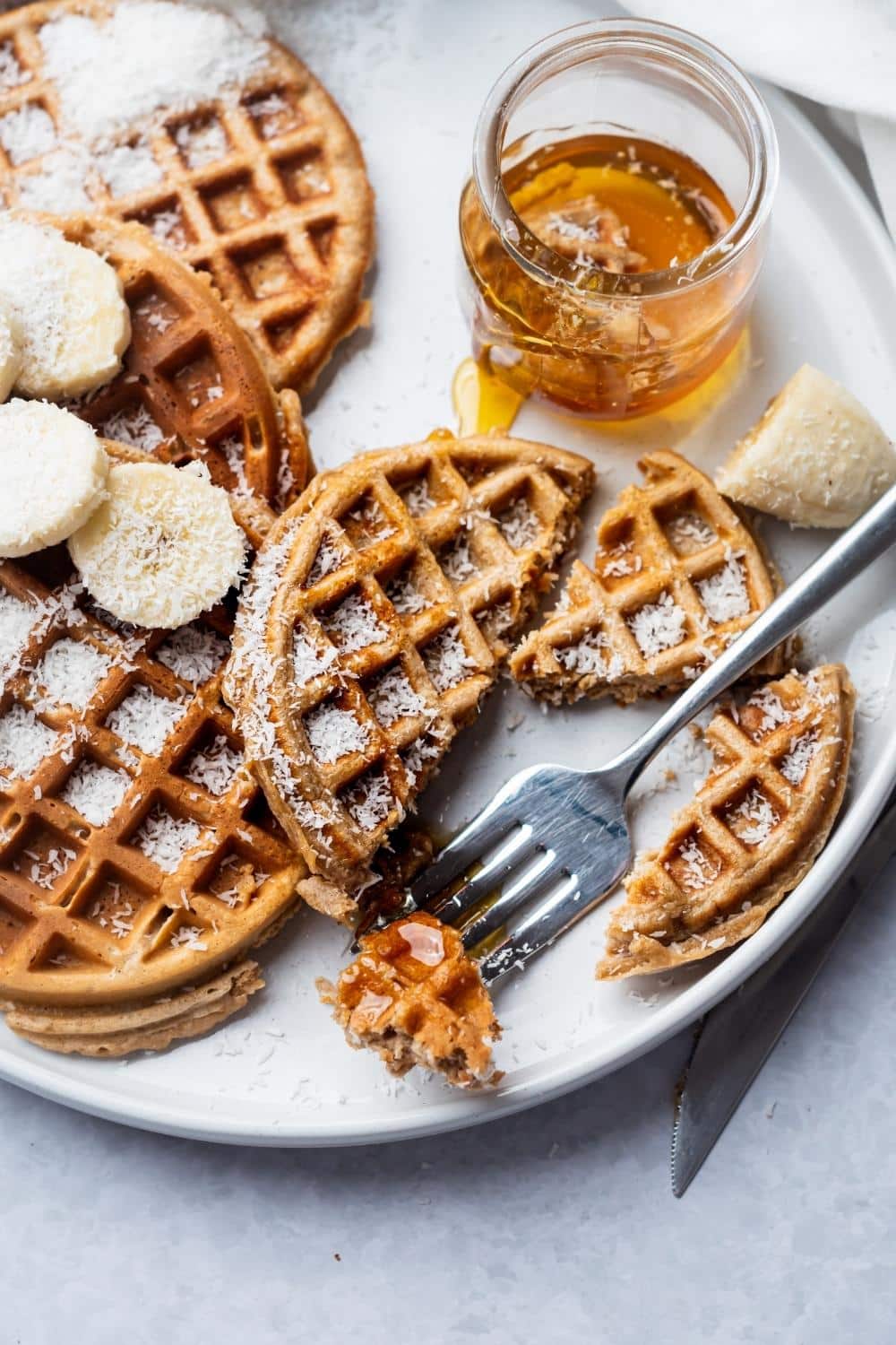 A protein waffle cut into pieces, and parts of three whole protein waffles on a white plate with a jar of syrup on it.