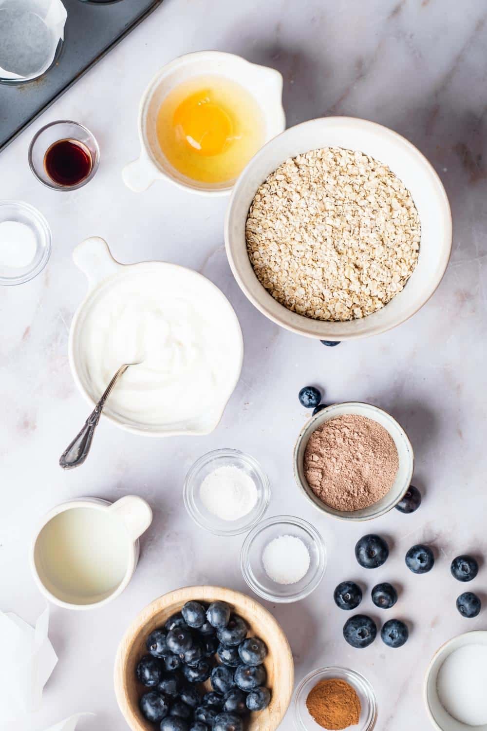 A bowl of oats, a bowl with an egg in it, a bowl of yogurt, a bowl of blueberries, and a bowl of protein powder all on a white counter.