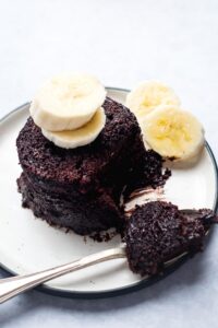 A chocolate protein mug cake on a white play with two slices of banana Anna, two slices of banana next week, and a spoon next to it with the mug cake on it.