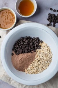 A white bowl with chocolate chips, protein powder, and oats in it. Behind that is a bowl of honey and peanut butter.