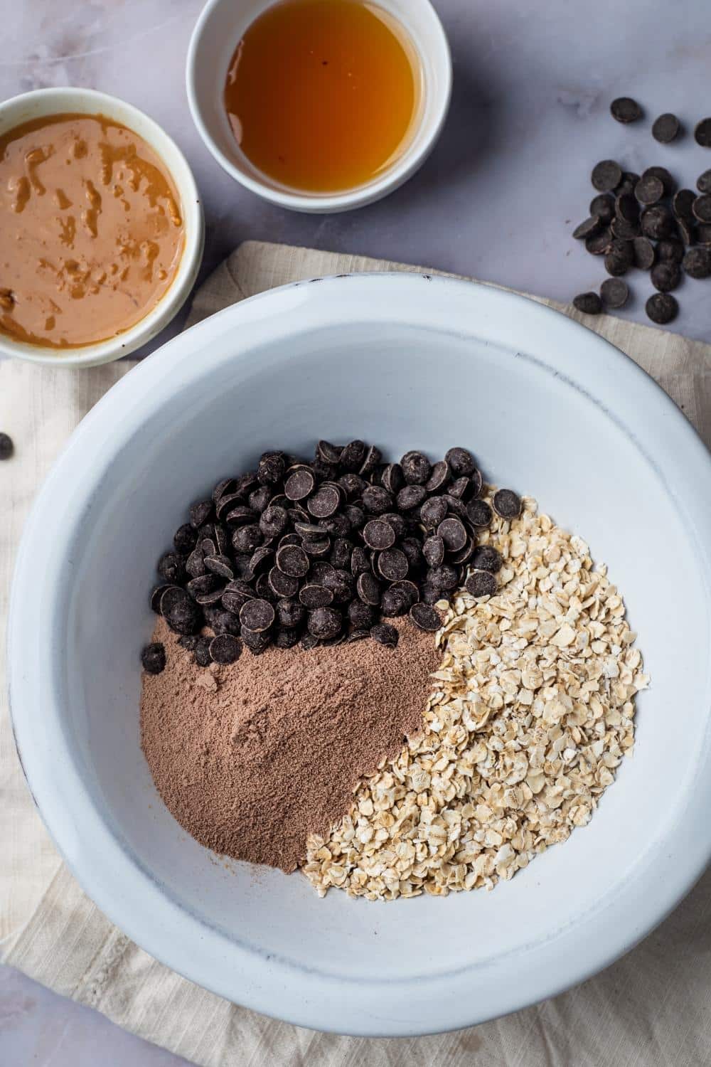 A white bowl with chocolate chips, protein powder, and oats in it. Behind that is a bowl of honey and peanut butter.