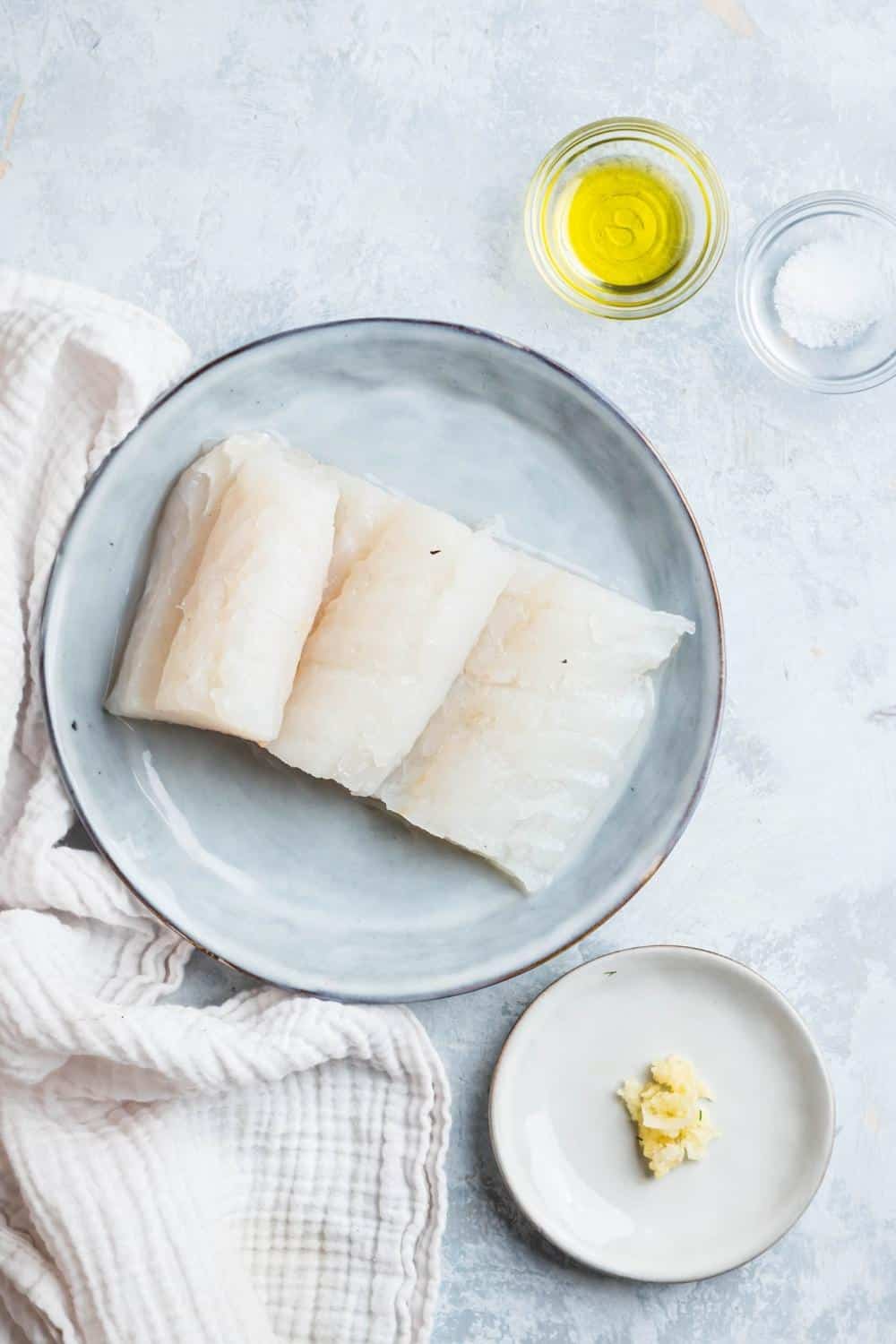 Halibut fillets on a grey plate, a bowl of olive oil, and a bowl of salt on a white counter.