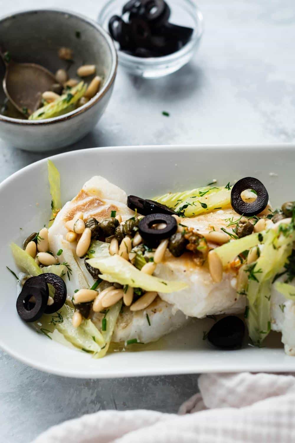 Black olives and pine nuts on top of halibut fillets on a white plate.