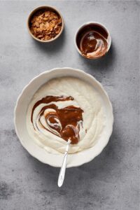 A bowl of mixed coconut milk, heavy cream, and streaks of almond butter. Behind it is a bowl of toasted almonds.