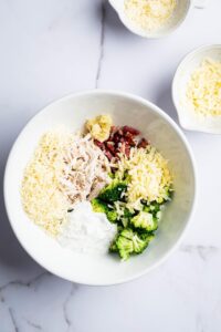 A white bowl filled with broccoli, bacon, shredded chicken, ranch dressing, and shredded cheese.
