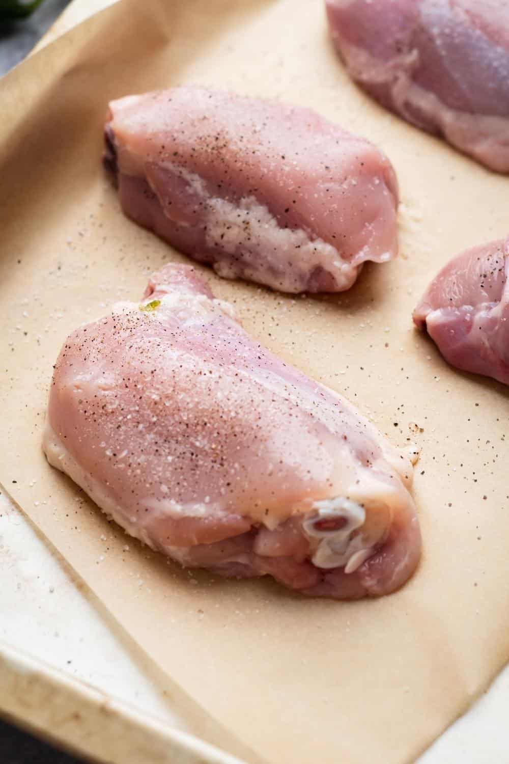 Raw chicken thighs with salt and pepper on top on a piece of parchment paper on a baking tray.