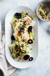 Black olives, celery, and pine nuts on top of halibut fillets on a white plate.